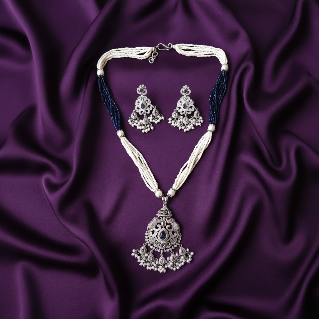A traditional Indian jewelry set featuring a silver-toned pendant necklace and matching earrings with white pearl-like strands and dark blue beaded accents, displayed on a draped purple satin fabric.