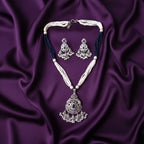 A traditional Indian jewelry set featuring a silver-toned pendant necklace and matching earrings with white pearl-like strands and dark blue beaded accents, displayed on a draped purple satin fabric.